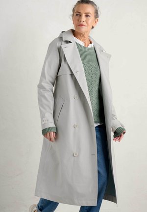 Manteau trench gris clair avec des boutons croisés, un grand col et des poches latérales. Porté sur un pull texturé vert, complété par un jean bleu.