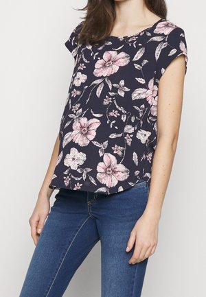 Mujer con blusa de manga corta azul marino con flores rosas y blancas combinada con jeans azules sobre un fondo liso.