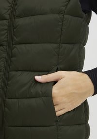 Tamnozeleni pufasti prsluk s kariranim uzorkom, bočnim džepovima i prednjim patentnim zatvaračem. Uvećani prikaz ruke koja se odmara u džepu ističe detalje dizajna.