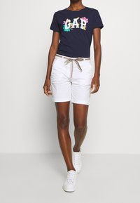 Osoba, která kráčí v bílých zkrácených kalhotách s proužkovým páskem, tmavě modrém květinovém tričku s logem GAP a bílých teniskách na jednobarevném pozadí.