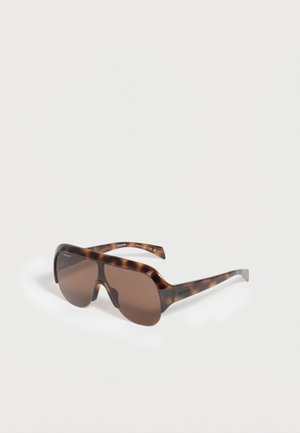 Nadmerné štítové slnečné okuliare z korytnačieho vzoru s hnedými prechodovými šošovkami a logom Gucci na ramenách, ležiace na bielej ploche.