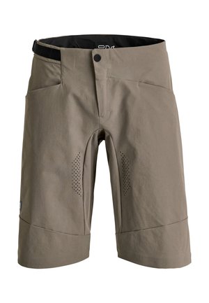 Mann trägt beiges langärmeliges Hemd, braune Shorts, schwarze Knieschoner, weiße Radsportschuhe, schwarzen Helm und Schutzbrille, steht mit Händen in den Taschen.