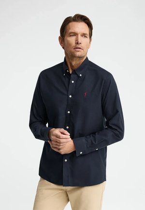 Hombre vistiendo una camisa azul marino abotonada con un pequeño logo rojo, pantalones beige, de pie contra un fondo claro y liso.