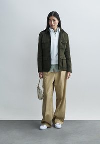 Πρασινοελιά μπουφάν με πολλαπλές τσέπες, λευκό φούτερ, ριγέ πουκάμισο, μπεζ παντελόνι φαρδιάς γραμμής και λευκά αθλητικά παπούτσια· κρεμ τσάντα.