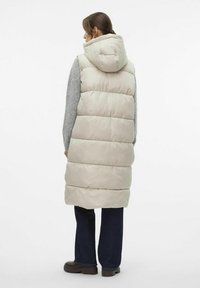 Gilet matelassé beige avec capuche, au design matelassé et à la texture extérieure lisse. Porté par-dessus un pull gris et associé à un jean foncé et des bottes marron.