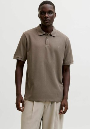 Ung mann med mørk hud som har på seg en kortermet brun poloshirt og lysebeige bukser med folder, stående mot en ensfarget bakgrunn.