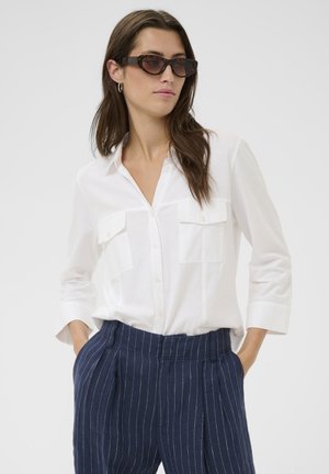 Woman wearing tortoiseshell sunglasses, white button-up shirt with chest pockets, and blue pinstriped pants, hands in pockets, looking sideways.