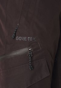 Gros plan sur un tissu GORE-TEX marron foncé avec deux poches zippées dotées de cordons noirs et blancs.