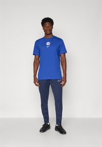 Blå Nike T-shirt med Chelsea FC-logo, parret med marineblå træningsbukser med et hvidt Nike-swoosh. Mørket fodtøj fuldender outfittet.
