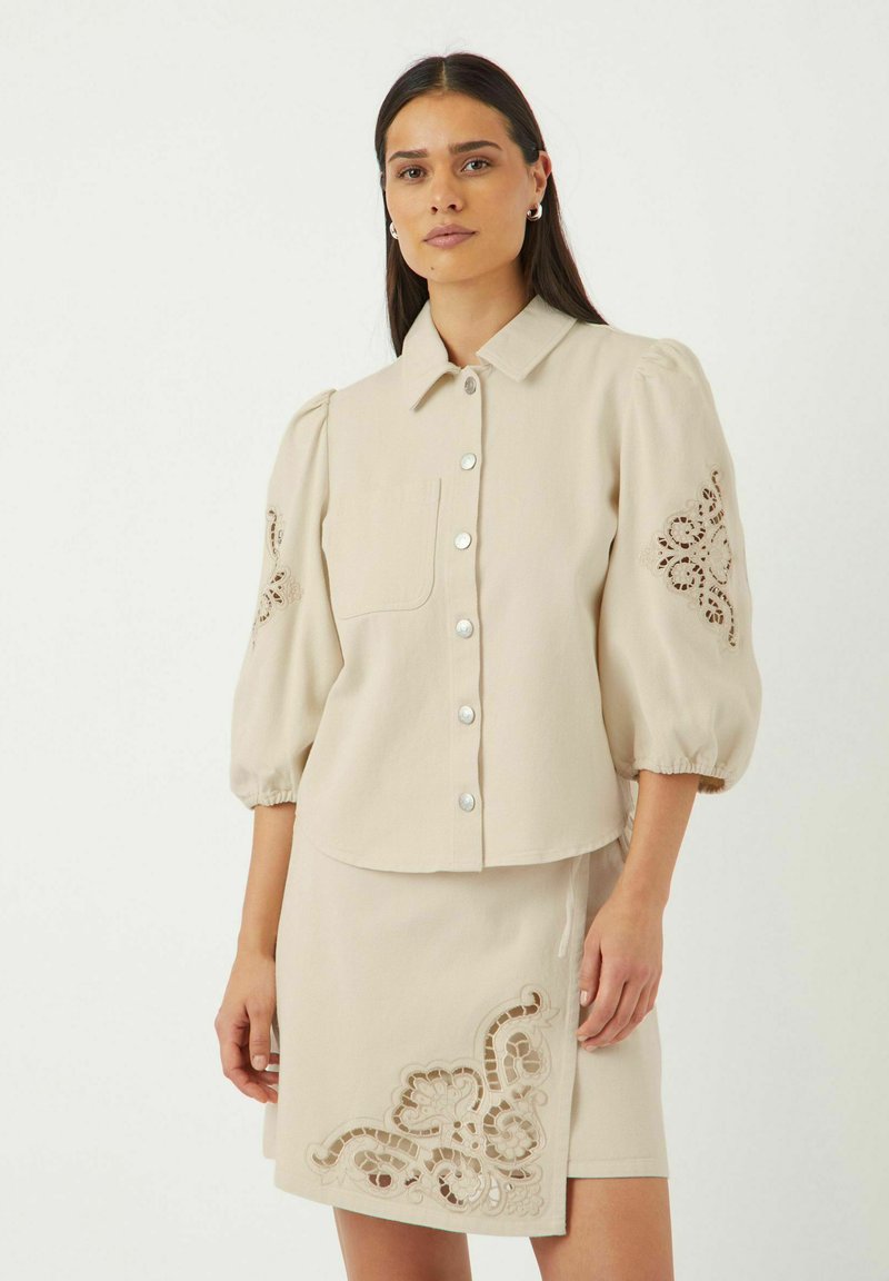 Beige bluse med puffede ærmer, knaplukning foran og lomme. Har broderede detaljer på ærmerne. Sammen med matchende beige nederdel med udsmykket mønster.