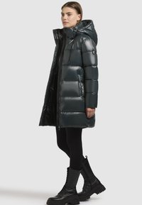 Polstret parka i mørkegrøn med blank overflade, der har hætte, lynlås lukning og sidelommer, båret over sorte leggings og militærstøvler.