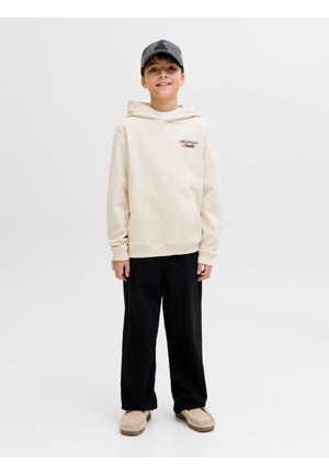 Chlapec stojaci a usmievajúci sa, oblečený v béžovej mikine s nápisom "Originals Studio", čiernych nohaviciach, béžových topánkach a sivej čiapke na bielom pozadí.