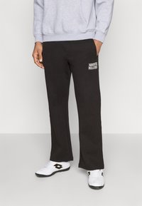 Crne trenirke od mekog materijala s ravnim krojem. Ima bočne džepove i bijeli natpis na donjem lijevom dijelu. Uparene su s bijelim sportskim cipelama.