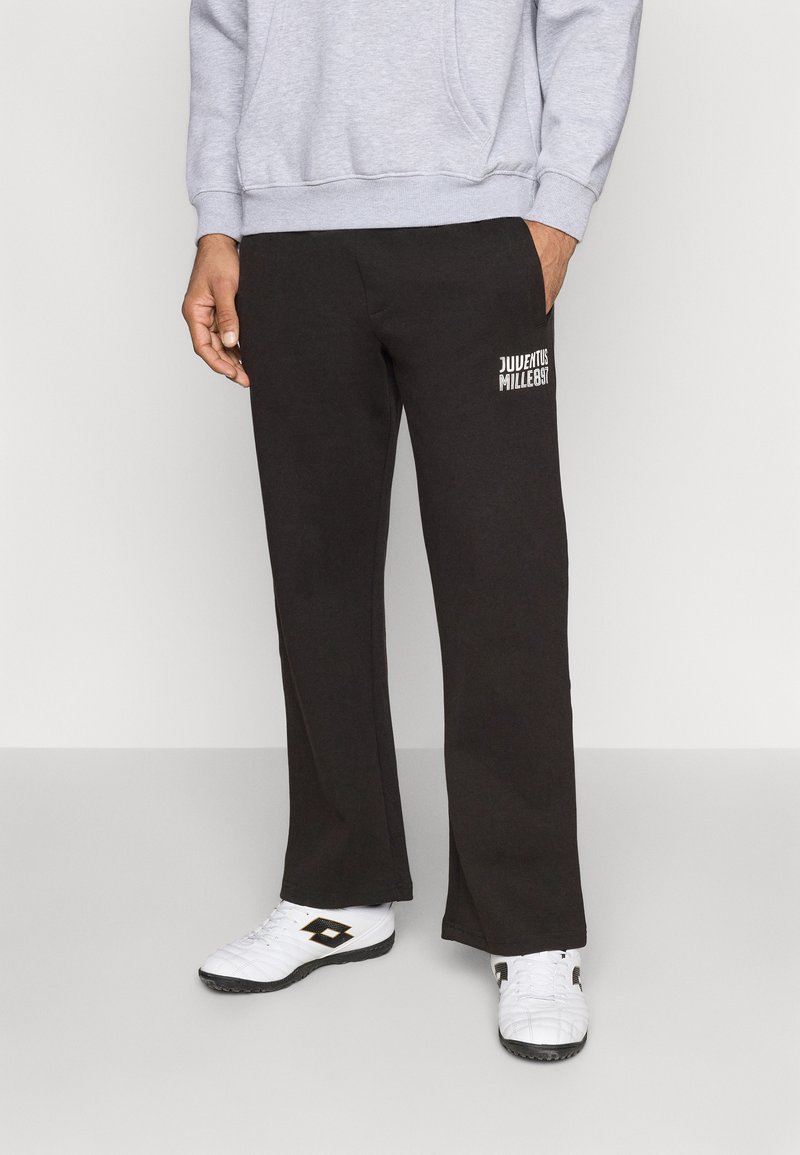 Crne trenirke od mekog materijala s ravnim krojem. Ima bočne džepove i bijeli natpis na donjem lijevom dijelu. Uparene su s bijelim sportskim cipelama.