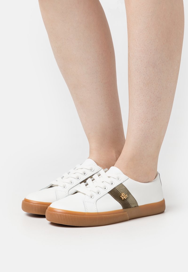 Baskets blanches avec semelles en gomme marron et bandes latérales vert foncé avec logo doré, portées pieds nus sur fond blanc.