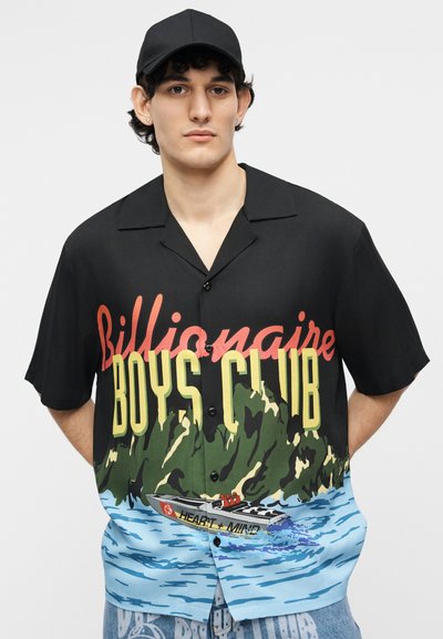 Fiatal férfi fekete sapkával és fekete, gombos inggel, melyen a "Billionaire Boys Club" felirat és színes hajó- és vízgrafika látható.