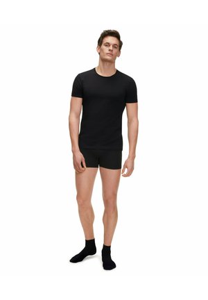 Black fitted t-shirt and boxer briefs, made of smooth fabric. Model stands in black socks, showcasing a simple, monochromatic outfit.