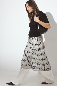 Melnā caurspīdīgā mežģīņu pārklājuma svārki ar ziedu rakstu, valkāti virs baltām plašām biksēm, un kombinēti ar pieguļošu melnu topu un melnu somu.