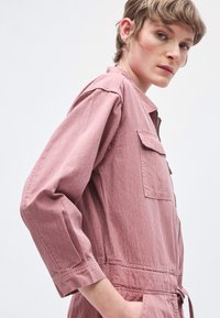 Rózsaszín pamut overál hosszú ujjakkal, elülső zsebekkel és megkötős derékkal. Sima textúrájú, enyhén strukturált kialakítás.