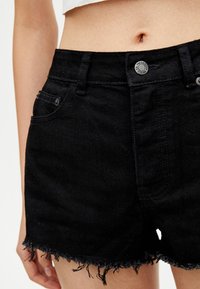 Pantalones cortos de mezclilla negra con dobladillo deshilachado, diseño de cintura alta, estilo de cinco bolsillos y cierre con botón de metal. Tejido texturizado con áreas desgastadas.