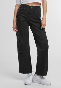 Pantaloni cargo neri con design a gamba dritta, numerose tasche laterali e chiusura frontale con bottone, abbinati a sneakers bianche.