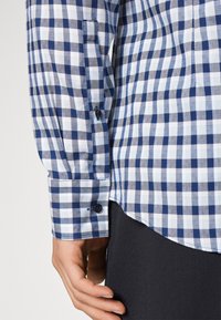 Main portant une chemise à manches longues à carreaux bleus et blancs, retroussée avec des boutons foncés, reposant à côté d'un pantalon noir.
