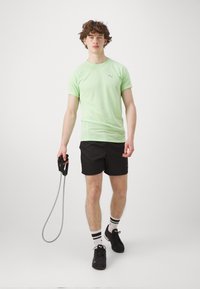 Camisa atlética verde claro de manga corta con un logo gris, combinada con pantalones cortos negros. La persona sostiene una cuerda para saltar en una mano.