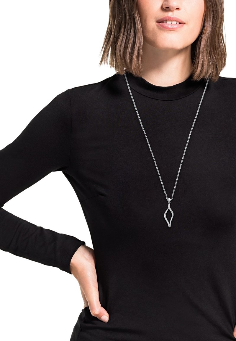 Zilveren hanger met een bladvormig ontwerp, hangend aan een fijne ketting, gedragen boven een hoge hals zwarte lange mouwen shirt.