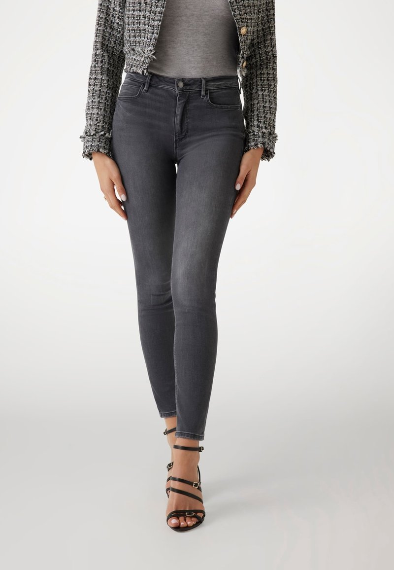 Femme portant un jean skinny gris foncé taille haute, une veste courte texturée et des sandales noires à talons hauts à brides, debout avec les mains détendues.