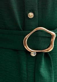 Zielony materiałowy pasek z charakterystyczną, nieregularną złotą metalową klamrą. Dodatkowy srebrny guzik widoczny na materiale.