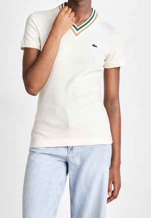 Person trägt ein cremefarbenes Polo mit kurzem V-Ausschnitt, grünen und beigen Streifen sowie hellblauen Jeans, Hand am Hals.