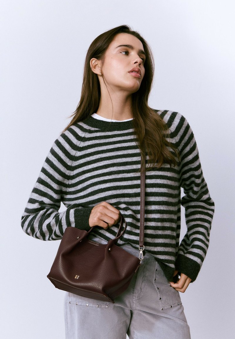 Suéter a rayas en gris y negro, sosteniendo un pequeño bolso de cuero burdeos con dos asas. Tela texturizada y sutiles acentos de hardware visibles.