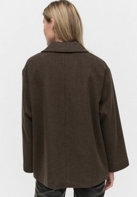 Bruine, gestructureerde jas met een brede kraag, verlaagde schouders en een rechte snit, voorzien van een rugventilatie en een ontspannen pasvorm.