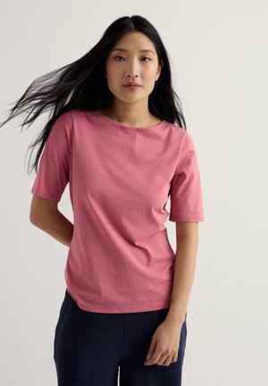 Ženska z dolgimi črnimi lasmi, oblečena v roza majico s kratkimi rokavi in temne hlače, stoji z eno roko za hrbtom.