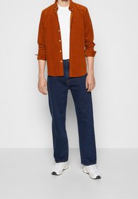 Rostfärgad kordflodsskjorta med knappar, bärs över en vit T-shirt, tillsammans med lösa blå jeans och vita sneakers. Enkel design, avslappnad passform.