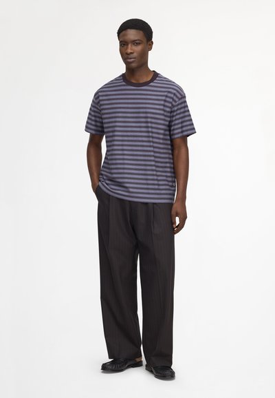 Jeune homme debout les mains dans les poches, portant un t-shirt rayé violet et noir, un pantalon large noir à fines rayures et des mocassins noirs.