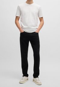 T-shirt bianca in cotone con maniche corte e un logo discreto sul lato sinistro del petto, abbinata a pantaloni neri e sneakers color crema.