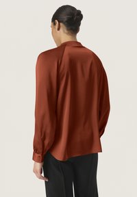 Blusa color ruggine in tessuto morbido, con una vestibilità comoda, maniche lunghe e polsini con bottone. Il design posteriore è semplice con un colletto minimale.