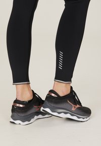 Mustat urheilukengät, joissa on teksturoitu pinta, ruusukultaiset yksityiskohdat ja harmaat kumipohjat. Jalassa tiukat mustat legginsit, joissa on heijastavia yksityiskohtia.