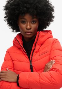 Rode pufferjack met een ritssluiting en capuchon, voorzien van een gewatteerde structuur. Merkenlogo op de borst. Warm ontwerp dat geschikt is voor koud weer.