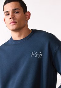 Sudadera azul marino de algodón, con un escote redondo y un logo blanco en el pecho. Textura suave, corte relajado.