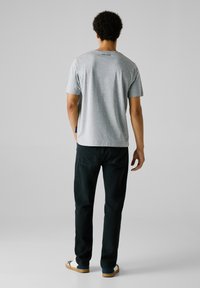 Camisa de algodón gris con mangas cortas, logo en la parte posterior, combinada con jeans oscuros. El modelo está de espaldas, usando zapatillas casuales con una suela estampada.