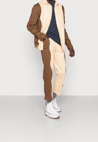 Brun och krämfärgad skjorta med knappar och ficka, matchad med bruna och krämfärgade byxor. Vita högre sneakers fullföljer outfiten.