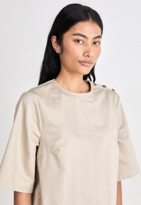 Camisola em mistura de algodão bege, com decote redondo, mangas curtas e botões decorativos no ombro; textura suave e design minimalista.