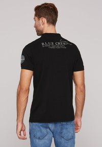 Polo shirt nera con maniche corte, colletto e stampe testuali sulla schiena. Il tessuto appare liscio con una leggera elasticità.