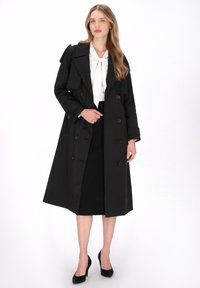 Zwarte dubbelgeknoopte trenchcoat met schouderepauletten, lange mouwen en een getailleerde taille, gecombineerd met een witte blouse en een zwarte rok.