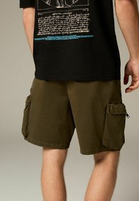 Shorts cargo kaki avec plusieurs poches latérales, une coupe décontractée et une texture de tissu lisse, portés avec un t-shirt noir à motif.