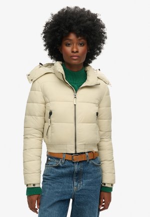 Beige verkürzte Puffjacke mit Kapuze, zwei vorderen Reißverschlusstaschen und elastischen Bündchen, getragen über einem grünen gerippten Pullover und blauen Jeans.
