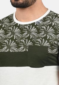 Camiseta verde y blanca con un patrón floral en la parte superior, con una sección verde sólida y un pequeño bolsillo en el pecho. Textura de tela suave.
