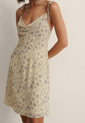 Żółta sukienka bez rękawów z fioletowym kwiatowym nadrukiem, zawiązywana na paski i z dekoltem typu komin, noszona przez osobę z rękami za plecami.